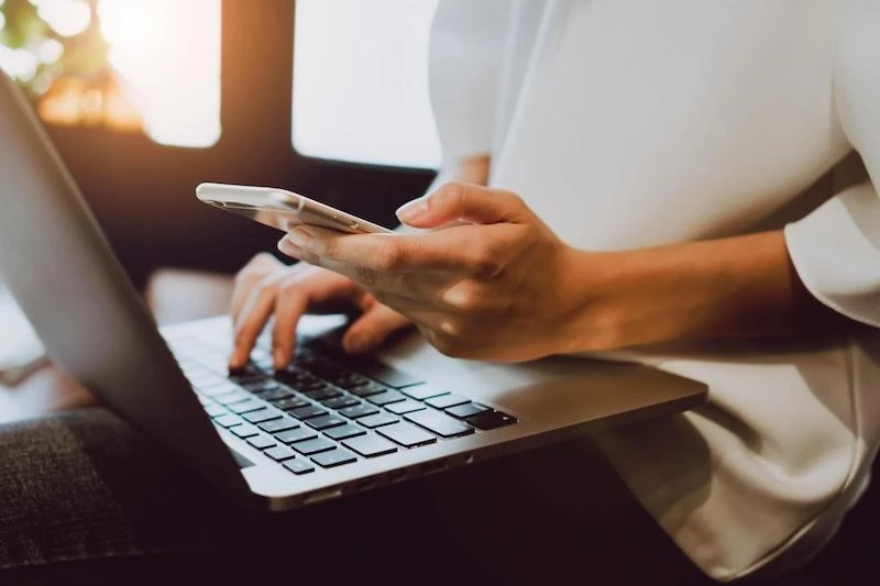 A woman typing in a laptop with a cellphone in the other hand